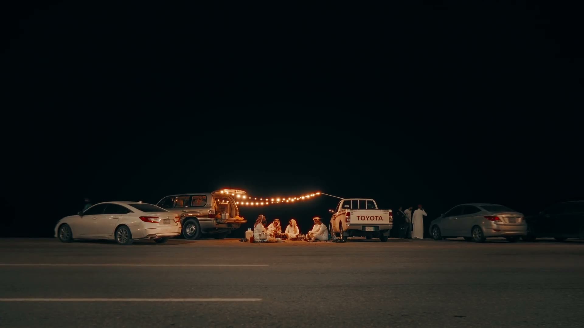 Night gathering lit by festoon lights between parked cars